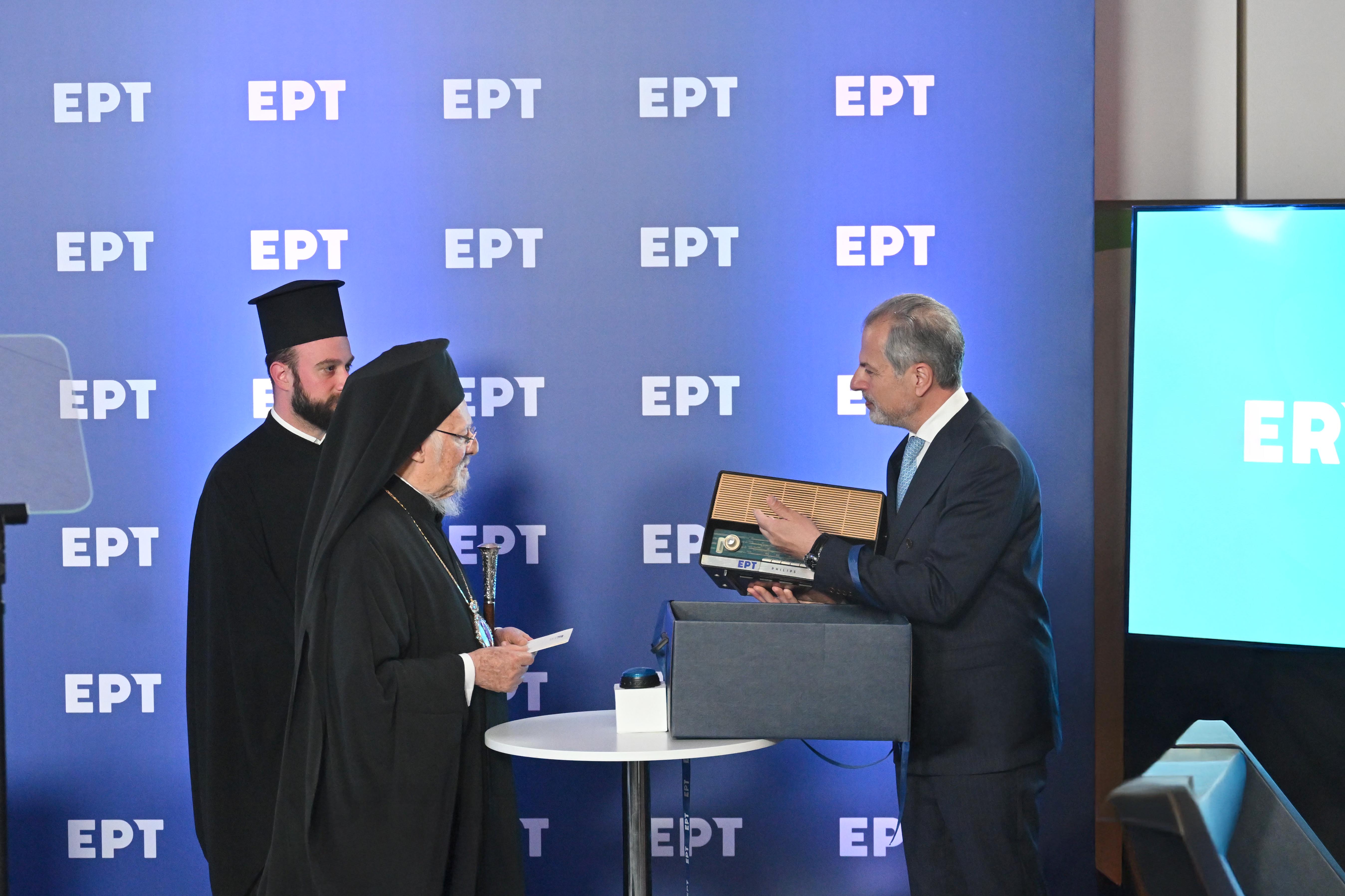 Ο Οικουμενικός Πατριάρχης Βαρθολομαίος και ο πρόεδρος της ΕΡΤ Γιάννης Παπαδόπουλος στέκονται δίπλα-δίπλα στο Ραδιομέγαρο, κρατώντας μια συλλεκτική συσκευή παλαιού ραδιοφώνου με ξύλινη επένδυση
