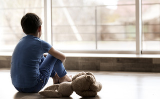 Παιδί λυπημένο κοιτάζει έξω από το παράθυρο έχοντας δίπλα του ένα λούτρινο